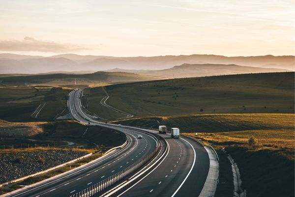 An elevated, expansive view of a winding highway cutting through rolling green hills and fields at sunset, with distant mountains and several semi-trucks traveling on the road.