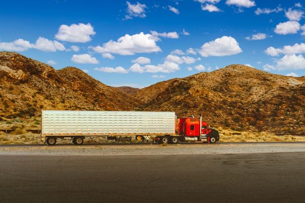 A vibrant red semi-truck pulling a white enclosed dry van trailer drives along a scenic highway through arid, mountainous terrain under a bright blue sky.
